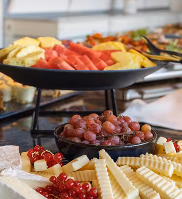 Assorterede frugter og oste vises på et buffetbord. Spredningen inkluderer skåret vandmelon, ananas, vindruer, forskellige oste og desserter arrangeret på sorte serveringsfade.