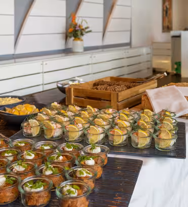 Buffetbord med et udvalg af mad, herunder små glaskrukker med forretter, et fad med skiveskåret brød og en skål med ostetern. Hvidt moderne design og et blomsterarrangement er synligt i baggrunden.