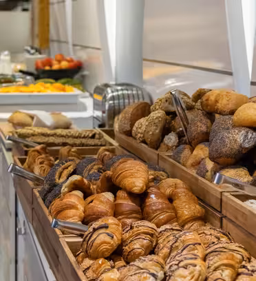 Et langt buffetbord viser et udvalg af brød og kager i træbakker, inklusive croissanter og rundstykker. I baggrunden er tallerkener med frugter og en brødrister synlige i moderne spisesteder.