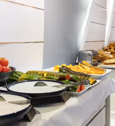 Et buffetbord med forskellige frugter, yoghurt og bagværk. Tallerkener med æbler, vandmelon og andre frugter er ved siden af skåle med yoghurt. Længere hen ad bordet står brød og kager på en hvid dug.