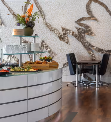 Et moderne spiseområde med et buet buffetbord med diverse madvarer og en grøn vase med blomster. Baggrunden har en stor abstrakt vægdekoration. Mørke stole og borde er arrangeret på en indbydende måde på et trægulv.