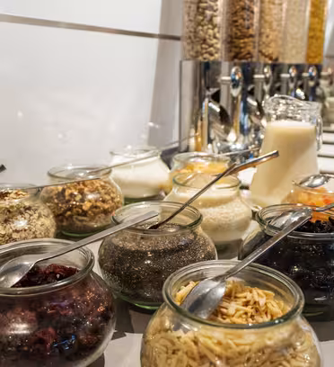 En morgenbuffet med glaskrukker fyldt med forskellige kornprodukter, nødder, tørret frugt og frø. En beholder med mælk er i baggrunden sammen med flere dispensere med flere korn. Skeer lægges i glassene.