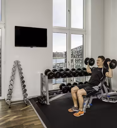 En person løfter håndvægte, mens han sidder på en bænk i et moderne fitnesscenter. Værelset har store vinduer med udsigt over byen og vandet, og der er et stativ med håndvægte ved siden af et væghængt tv.