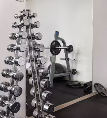 Et motionsrum med et stativ af sølvhåndvægte til venstre, en bænkpres med vægtskiver og en vægtstang i baggrunden og diverse gymnastikudstyr som en foam roller på gulvet. Væggene er hvide, og gulvet er dækket af sorte måtter.