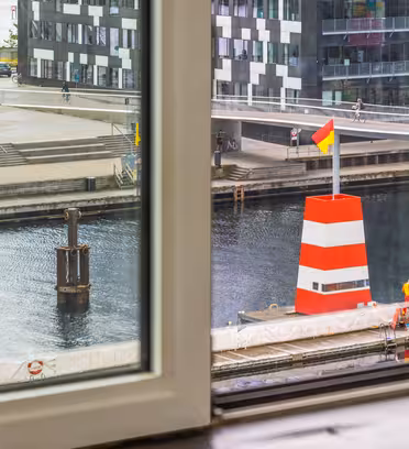 Udsigt gennem et vindue, der viser en rød og hvid bøje med et flag på toppen i en kanal. Moderne bygninger og en gangbro med mennesker og cykler er synlige i baggrunden.